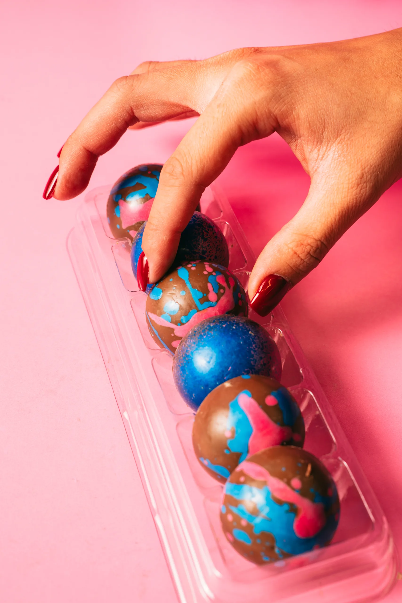 A hand lifting a chocolate sphere dessert from a clear tray on a pink backdrop