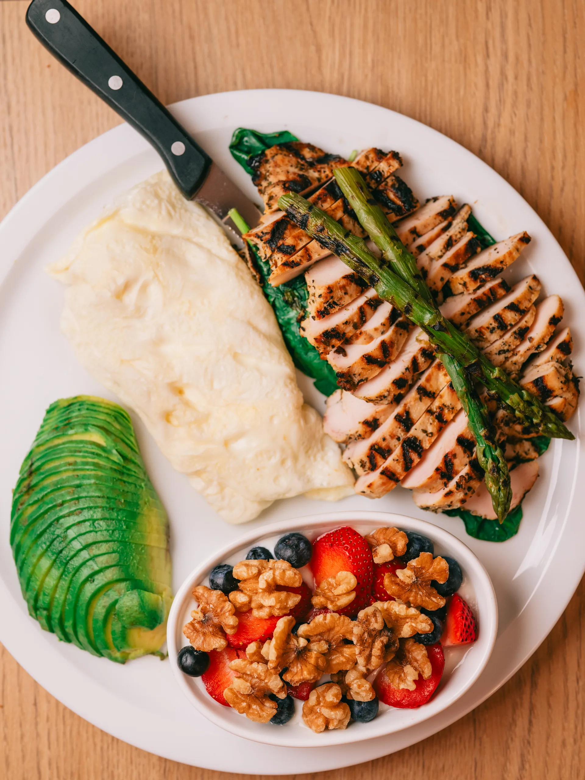 A keto breakfast plate of grilled chicken and asparagus, an egg white omelette, sliced avocado and a berry and walnut bowl