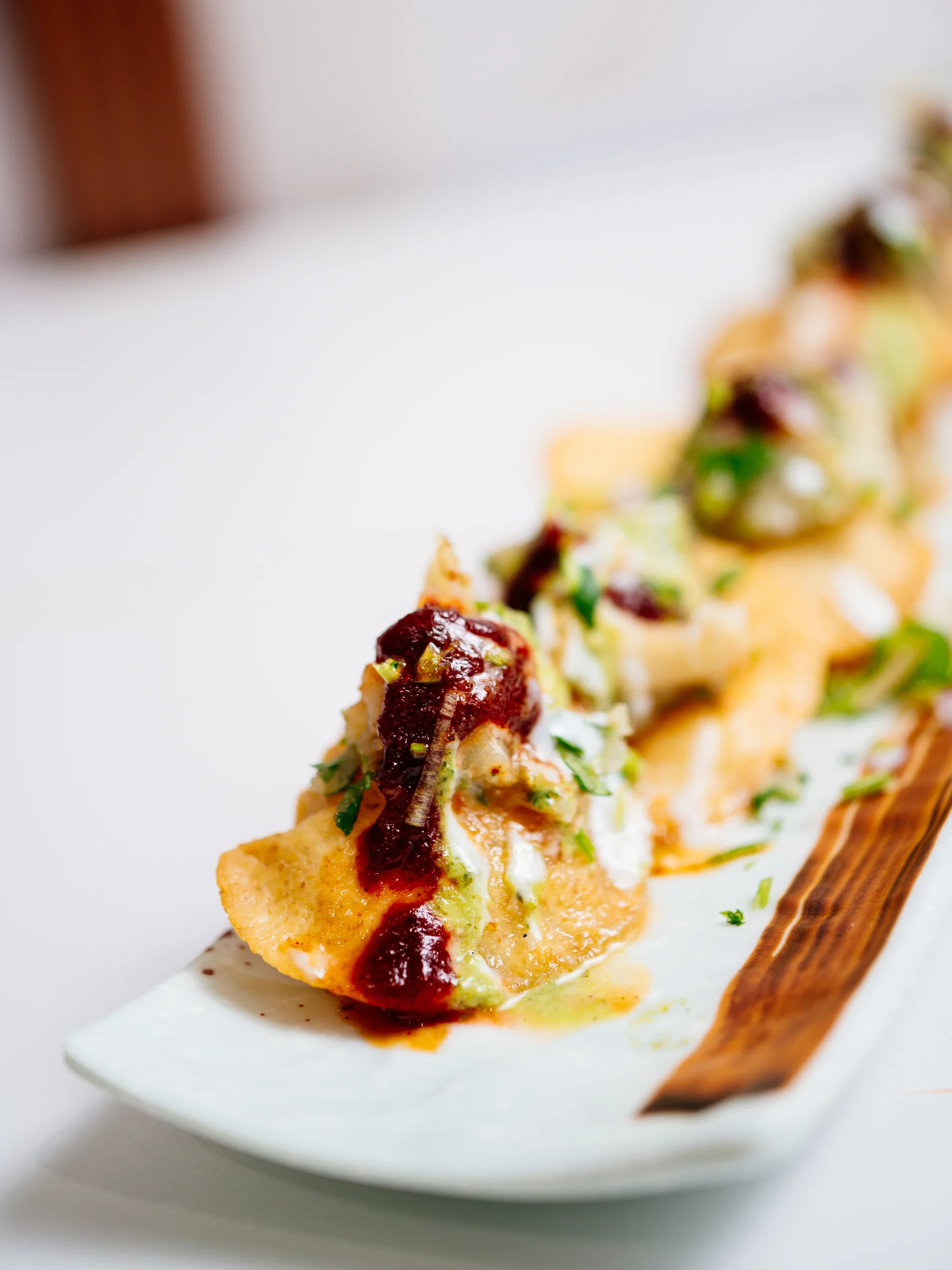 A row of chaat-topped samosas with tamarind, mint chutney and herbs on a long white plate