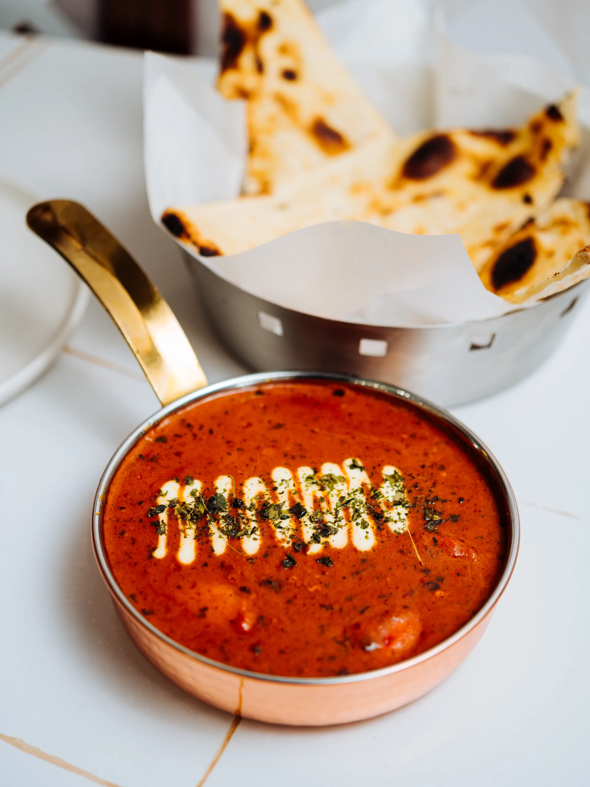 A copper karahi of creamy tomato curry drizzled with cream and herbs, beside a bowl of charred naan