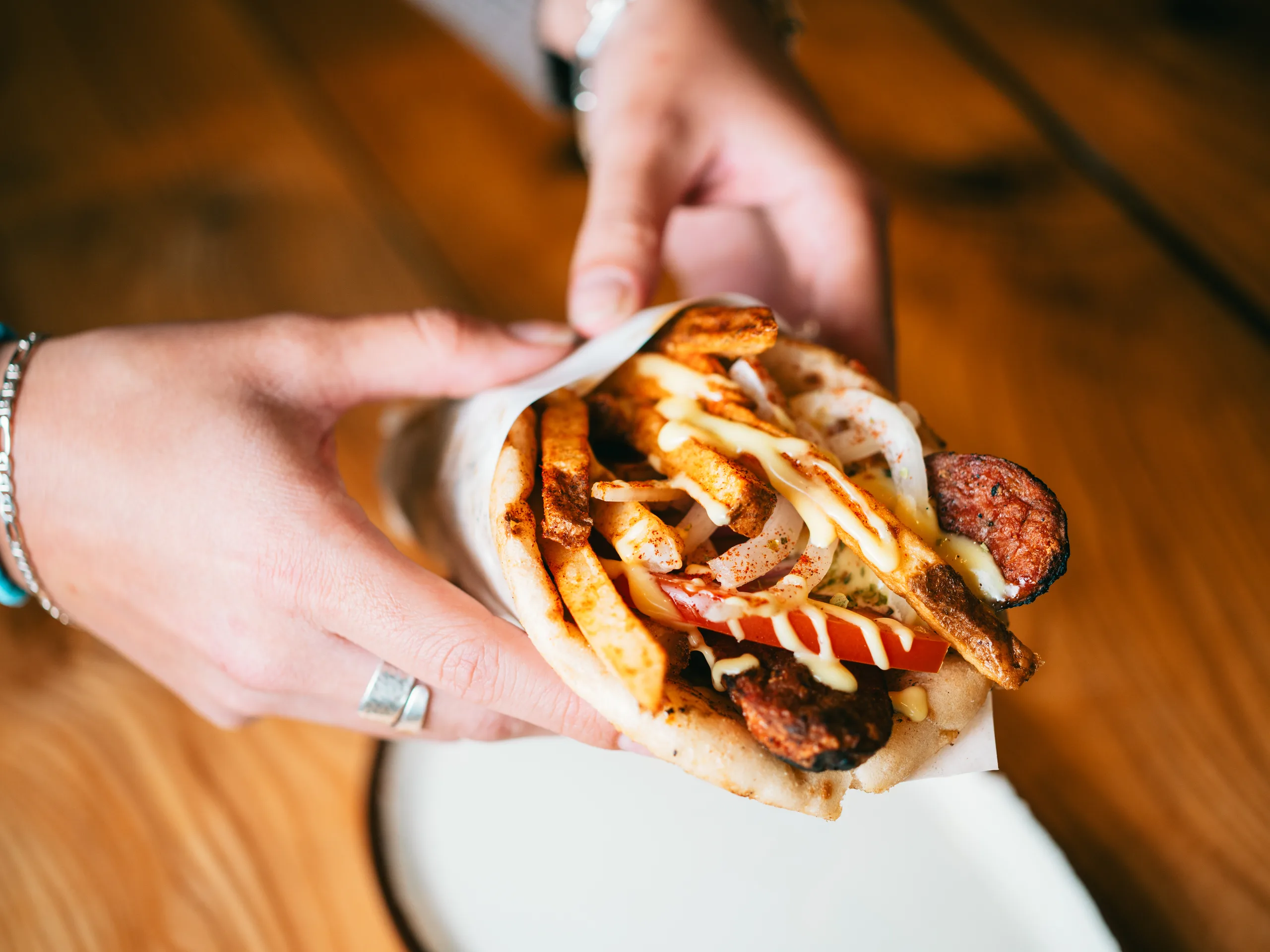 Hands holding a pita wrap filled with grilled meat, fries, tomato and garlic sauce over a wooden table